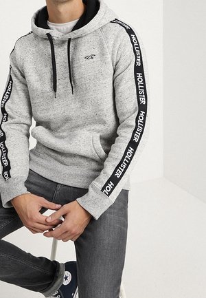 Mann trägt hellgrauen Hollister-Hoodie mit schwarzen Kordeln und Logostreifen an den Ärmeln, dunkle Jeans und marineblaue Sneakers, sitzt auf einem Hocker.