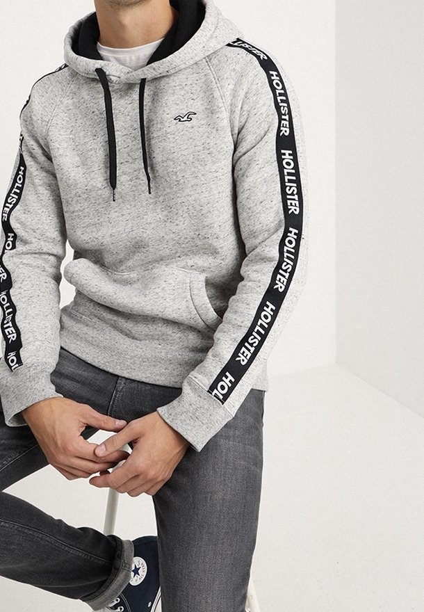 Homme portant un sweat à capuche Hollister gris clair avec des cordons noirs et des bandes logos sur les manches, jean foncé et baskets bleu marine, assis sur un tabouret.