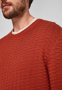 Sweter o rustykalnym kolorze rdzy z fakturą i prążkowanym wzorem, okrągłym dekoltem oraz kontrastowym kremowym wewnętrznym kołnierzem widocznym. Zbliżenie na detale tkaniny.