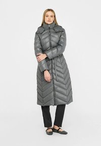 Grauer langer Steppmantel mit hohem Kragen, Chevron-Muster, Reißverschluss vorne und zwei Taschen. Das Modell trägt schwarze Hosen und Schuhe.