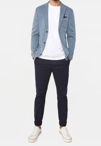 Man med ljusblå strukturerad kavaj, vit t-shirt, mörknavy byxor och vita sneakers som står med händerna i fickorna.