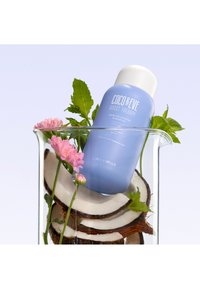 Blå schampoflaska med vit lock, omgiven av kokosbitar, rosa blommor och myntablad i en glasbehållare.