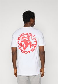 White cotton t-shirt featuring a large red graphic of a cartoon figure playing football, with "ADIDAS FOOTBALL" text surrounding the design.