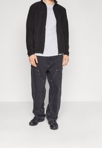 Chaqueta de forro polar negra con cuello alto, camiseta gris debajo, pantalones vaqueros grises oscuros holgados con cremalleras y zapatos negros con textura.