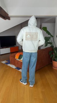 Osoba w białej bluzie z kapturem z napisem "WORLDWIDE '85" i niebieskich jeansach stoi w salonie z brązową kanapą i dużą doniczką z rośliną.