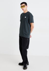 Dark green cotton t-shirt with a crew neck, paired with black loose-fitting jeans. Yellow-tinted sunglasses and black lace-up shoes visible.
