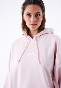 Felpa oversize rosa in morbido tessuto, dotata di tasca frontale, cappuccio con cordino e logo ricamato bianco sul petto.