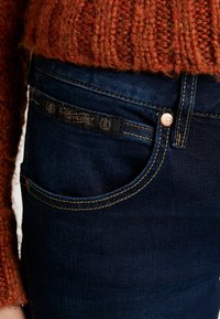 Denimjeans i djupblått med kontrasterande guldstickningar; har en etikett på bakfickan och en kopparknapp för stängning.