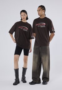 Brune oversized t-shirts med "OFFLINE FOR PRIVATE REASONS" tryk, båret med sorte cykelsorts og sorte støvler til knæene. Modellen har slidte jeans på.