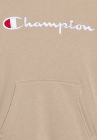 Hoodie beige realizzato in tessuto morbido, con logo "Champion" ricamato bianco e una tasca frontale con dettagli di cucitura visibili.
