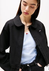 Sort lynlåstrøje med rundet krave og diskret logo. Båret over en lyseblå top med blonderkant og lodrette striber.