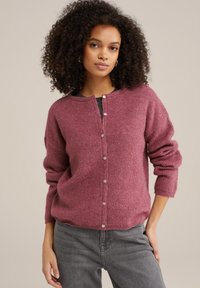 Trikotažna jopica v mehki, strukturirani sivkasti barvi z okroglim izrezom, sprednjimi gumbi in sproščenim krojem. Kombinirano z sivo naravnimi hlačami.