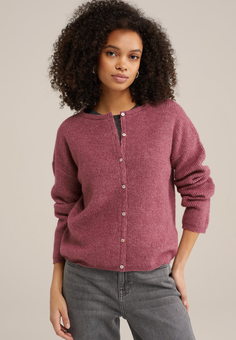 Trikotažna jopica v mehki, strukturirani sivkasti barvi z okroglim izrezom, sprednjimi gumbi in sproščenim krojem. Kombinirano z sivo naravnimi hlačami.