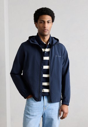 Man wearing navy hooded jacket over black and white striped polo shirt with light blue jeans against plain light background.