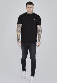 Svart t-shirt med kort ärm och rund halsringning, prydd med en liten logotyp på bröstet, tillsammans med mörka jeans och vita sneakers.