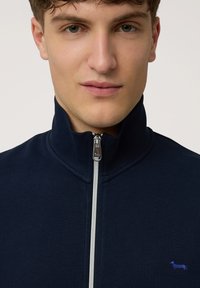 Chaqueta de forro polar azul marino con cremallera, cuello alto, y pequeño bordado de un perro azul en el lado izquierdo del pecho. Textura suave.