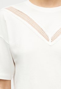 Haut blanc à manches courtes avec un détail en dentelle en forme de V sur la poitrine supérieure et les épaules, sur une peau claire.