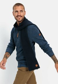 Navy knitted sweater with a zip collar, paired with a ribbed dark blue scarf. Knit texture and a logo patch are present on the sleeve.