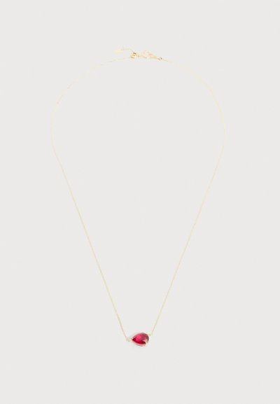 Λεπτό χρυσό κολιέ με μια μικρή καρδιόσχημη κόκκινη πέτρα ως μενταγιόν, τοποθετημένο στο κέντρο σε ανοιχτό γκρι φόντο.