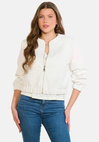 Teksturēta balta bombera jaka ar rāvējslēdzēju priekšpusē, ribotām aprocēm un apakšu. Valkāta pār gaišu topu, kopā ar džinsa biksēm.