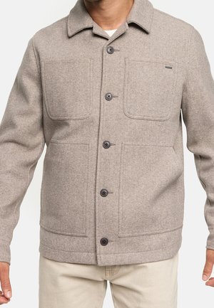 Mężczyzna w beżowej wełnianej kurtce na guziki z czterema przednimi kieszeniami oraz w kremowych spodniach.