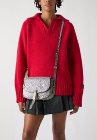 Rode gebreide trui met een kraag, gecombineerd met een zwart-wit gespikkelde crossbody tas met gouden hardware en een verstelbare schouderriem.