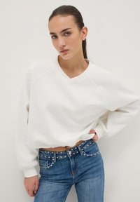 Sudadera blanca recortada con cuello en V y mangas raglán, combinada con vaqueros azules que tienen detalles con tachuelas en la cintura.