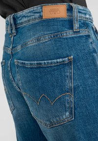 Jeans de mezclilla azul con una altura alta, que cuentan con un parche de cuero marrón, costuras naranjas y bolsillos traseros con un diseño curvado distintivo.