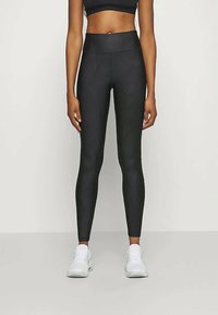 Melnas augstas vidukļa legingas ar fakturētu rakstu un elpojošu materiālu, apvienotas ar baltām sporta kurpēm neitrālā fonā.