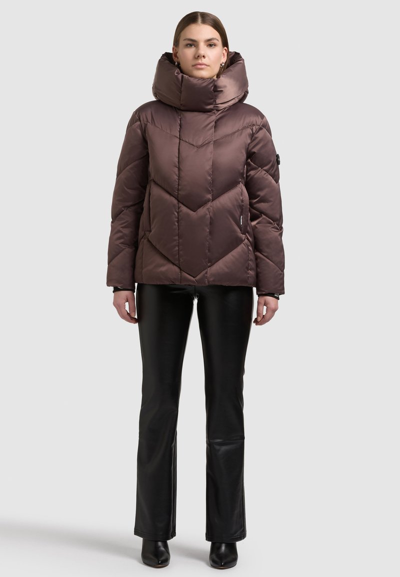 Bruine donzen jas met een hoge kraag en een chevronsteekpatroon. Zacht, glanzend materiaal; gecombineerd met zwarte strakke broek en enkellaarsjes.