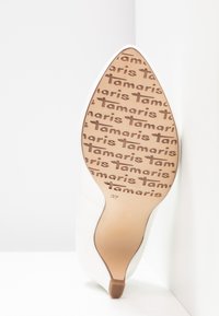 Escarpin blanc pointu levé contre un mur blanc, montrant la semelle avec le logo "Tamaris" répété et la taille 37.