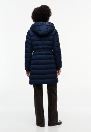 Personne aux cheveux bouclés portant un long manteau matelassé bleu marine avec capuche et un pantalon noir, debout de dos sur un fond uni.