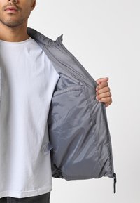 Chaqueta ligera gris con cierre de cremallera, que muestra un forro interno liso y brillante y un dobladillo ajustable. El exterior tiene una textura mínima y sin patrones.
