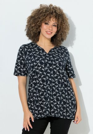 Kobieta o kręconych włosach ubrana w czarną bluzkę z krótkim rękawem z białymi abstrakcyjnymi wzorami linii, stoi na tle jednolitej jasnej ściany.