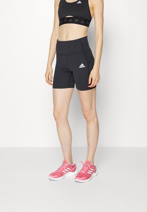 Frau trägt schwarzen Adidas Sport-BH und Shorts, kombiniert mit pinkfarbenen Adidas Sportschuhen, steht vor einem schlichten weißen Hintergrund.