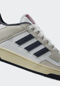Sneaker features a white leather upper with grey suede accents, three dark blue stripes, and a textured cream rubber sole. Laces are white.