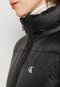 Black puffer jacket with a high collar, zip closure, and textured fabric; features a white logo on the left chest.