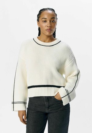 Maglione a coste bianco con strisce nere attorno al collo e all'orlo. Presenta maniche ampie e una silhouette corta. Abbinato a jeans scuri.