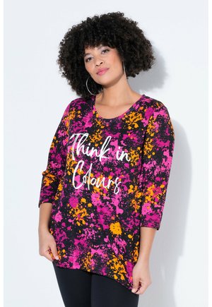 Kvinde med krøllet hår bærer store øreringe og en livlig top med sprøjtetryk i lilla, pink og orange med teksten "Think in Colours".
