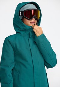 Veste d'hiver bleu canard avec une capuche, dotée d'une fermeture éclair sur le devant et d'un système de pression. Le porteur porte des lunettes de ski miroir et un bonnet tricoté.