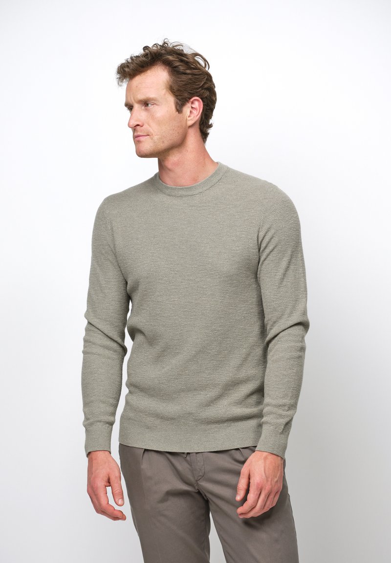 Maglione grigio chiaro con texture, collo a giro, maniche lunghe e polsini a coste, abbinato a pantaloni chiari.