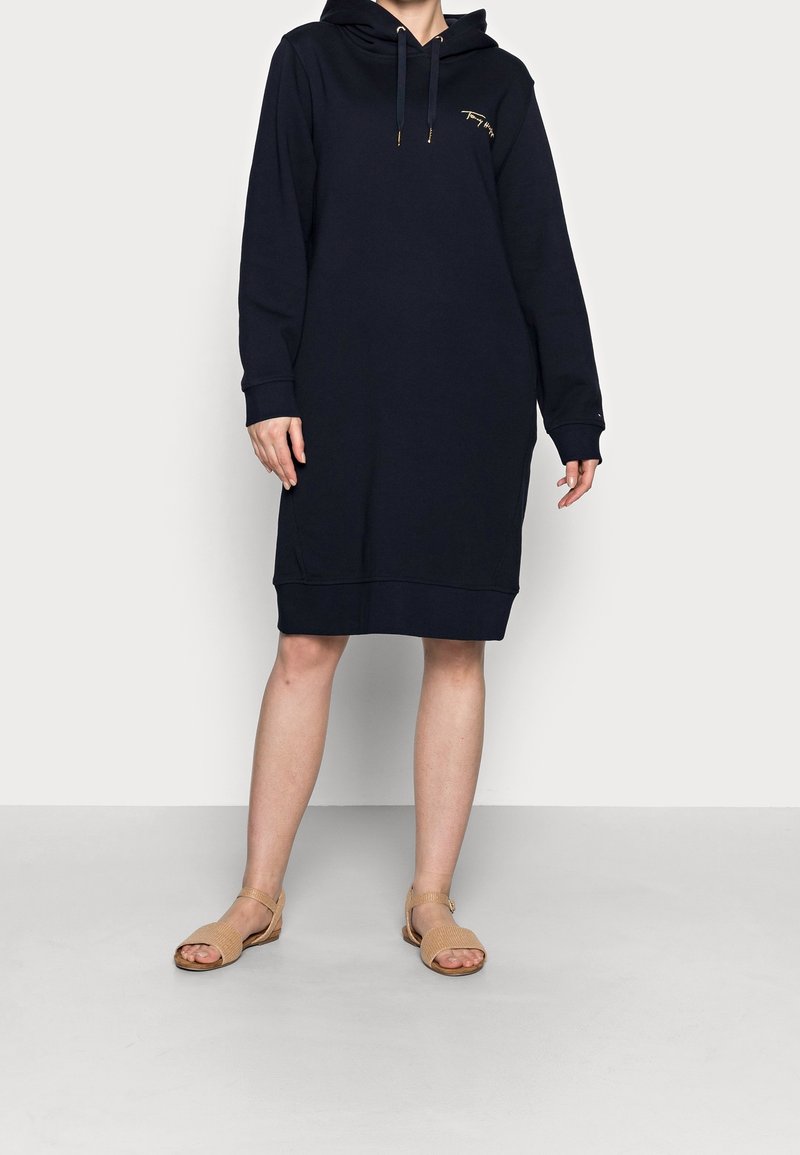 Marineblauwe hoodie-jurk met lange mouwen en een trekkoordkap. Voorzien van een gouden accentlogo, losse pasvorm en geribde zoom. Gemaakt om te combineren met beige sandalen.