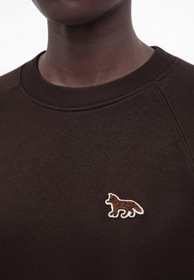 Κοντινό σε ένα άτομο που φοράει σκούρο καφέ φούτερ με ένα μικρό κεντημένο έμβλημα αλεπούς στο στήθος.