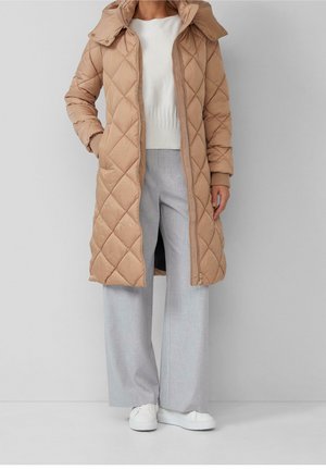 Femme portant un manteau matelassé beige mi-long, un pull blanc, un pantalon gris clair à larges jambes et des baskets blanches sur un fond gris uni.