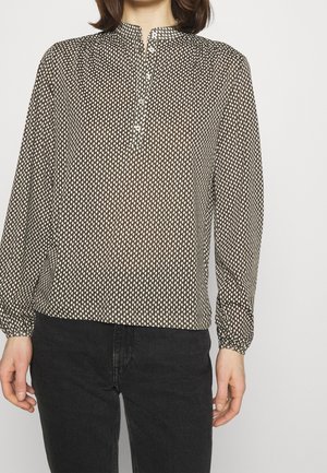 Femme portant un chemisier marron à manches longues avec un motif de gouttes blanches, col boutonné, et un jean noir sur un fond uni.