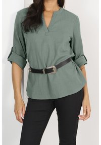 Camicia verde, leggera, a maniche lunghe con scollo a V e maniche arrotolate, abbinata a una cintura nera con fibbia in argento, indossata sopra pantaloni scuri.