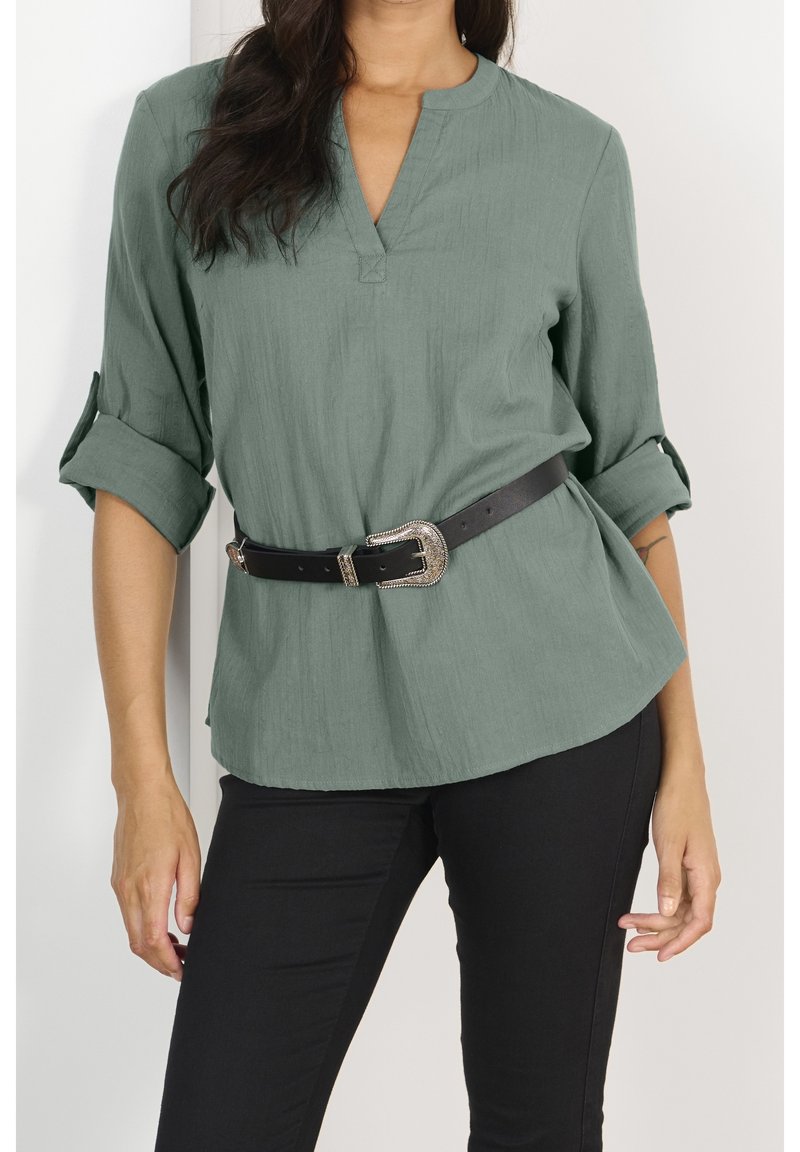 Camicia verde, leggera, a maniche lunghe con scollo a V e maniche arrotolate, abbinata a una cintura nera con fibbia in argento, indossata sopra pantaloni scuri.