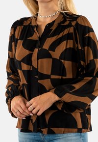 Blusa de estampado marrón y negro con botones en el frente, hombros fruncidos y mangas largas con puños elásticos. Textura de tela suave.