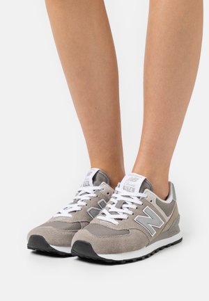 Persona che indossa sneaker New Balance 574 grigio chiaro con lacci bianchi, in piedi su una superficie bianca, che mostra la vista frontale e laterale delle scarpe.