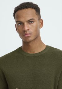 Maglione a maglia verde oliva con scollatura rotonda e tessuto testurizzato, caratterizzato da un design attillato che segue la forma del corpo.